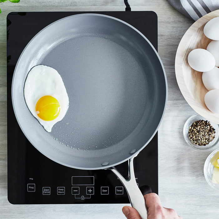 A hand holds a 12 Valencia Pro Ceramic Fry Pan by GreenPan on an induction cooktop, frying a sunny-side-up egg in PFAS-free cookware. Nearby are a bowl of white eggs, a small bowl of pepper, and a striped cloth.