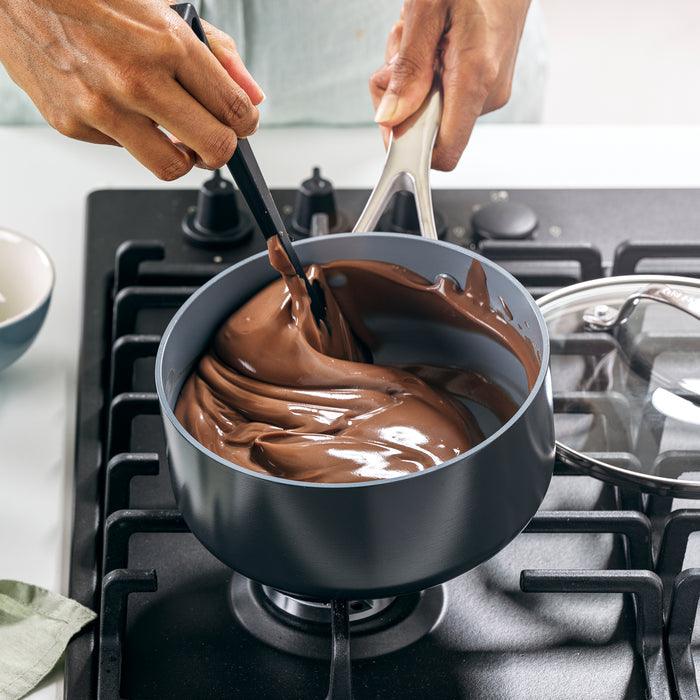 A person stirs melted chocolate in a GreenPan Valencia Pro Nonstick, Ceramic Sauce Pan with Lid, 2qt on the stovetop, enjoying PFAS-Free performance. Their hands skillfully manage the spoon while a glass lid sits nearby.