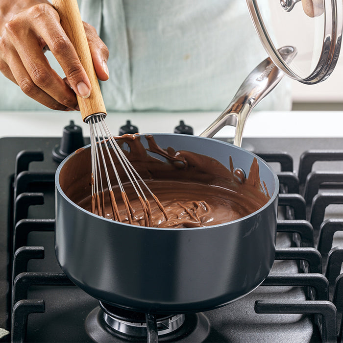 A person expertly stirs melted chocolate in a saucepan on a black stove using GreenPans Valencia Pro Nonstick, Ceramic Sauce Pan with Lid, 2qt. Their elegant hand and light-colored shirt enhance the cooking scene as they deftly manage the whisk and lid.