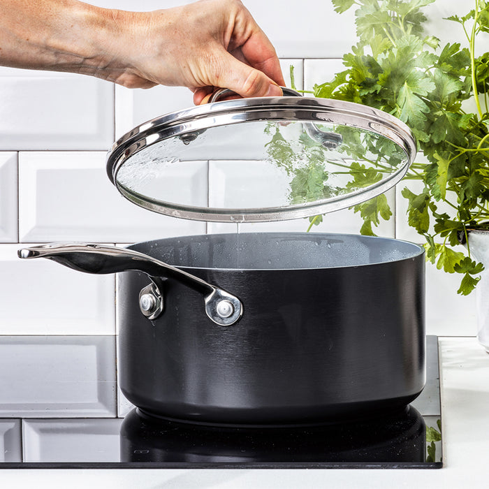 A hand lifts the glass lid of a 2qt Valencia Pro nonstick ceramic saucepan by GreenPan on a stovetop, releasing steam. Fresh cilantro contrasts against a white tiled backsplash. This PFAS-Free cookware ensures safe and effortless cooking with its durable ceramic nonstick coating.