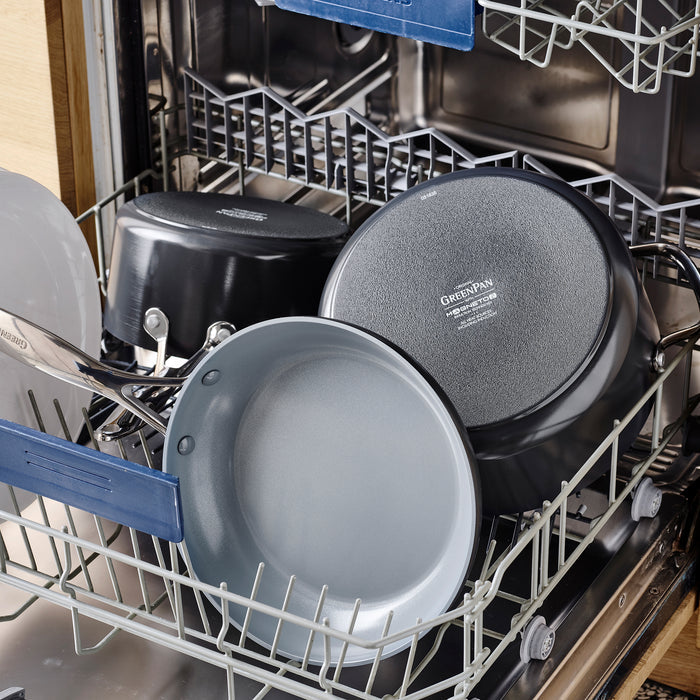 Open dishwasher with PFAS-Free GreenPan Valencia Pro cookware inside. Visible are a frying pan, a 2qt Valencia Pro Nonstick Ceramic Sauce Pan with Lid, and large plates. The extended racks neatly display the collection.