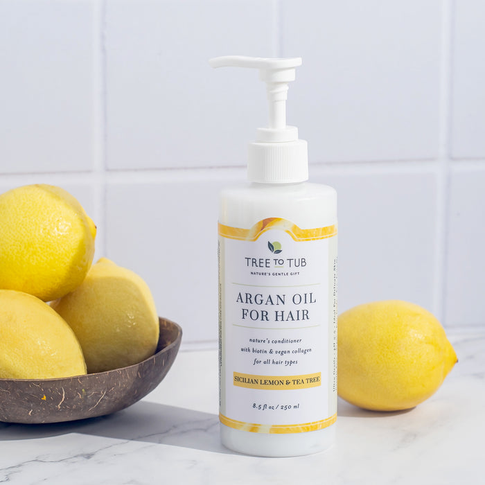 A bottle of Tree To Tubs Vegan Collagen & Biotin Conditioner for Fuller Stronger Hair sits on a white surface, labeled Sicilian Lemon and Tea Tree. It is accompanied by fresh lemons against pristine white tiles.