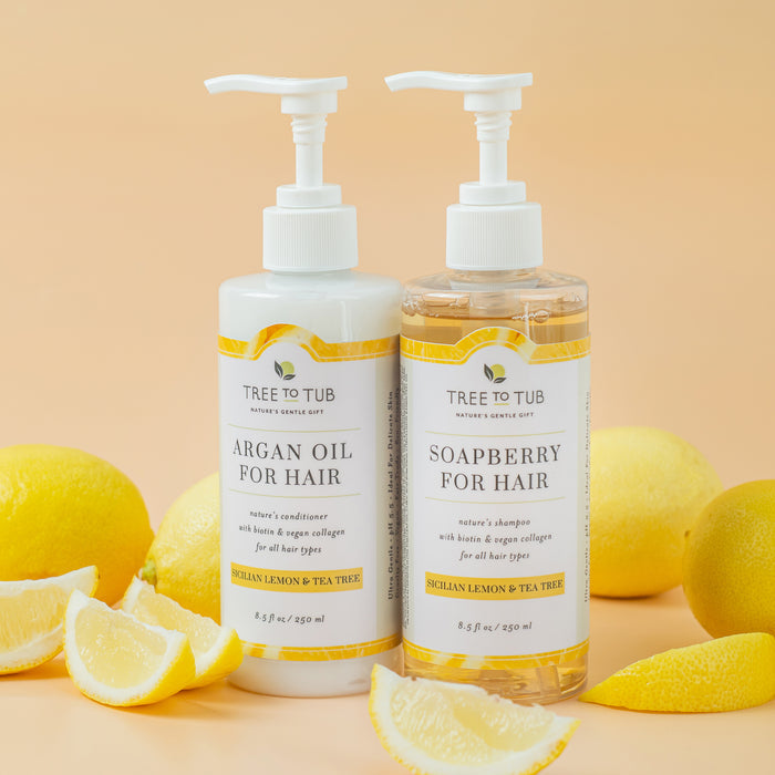 Two Tree To Tub hair care bottles, Argan Oil for Hair and Soapberry for Hair, are showcased with lemons. The Sicilian Lemon and Tea Tree bottles are set against a soft yellow background, enhancing the richness of each Vegan Collagen & Biotin Conditioner for Fuller Stronger Hair.
