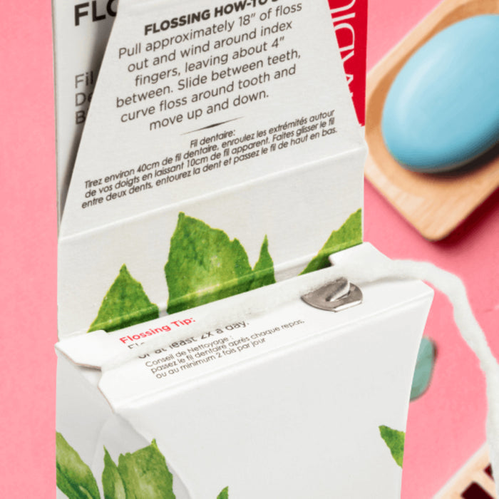 A close-up of an open Radius Vegan Floss box with instructions on the lid and organic floss pulled out, featuring a mint leaf design on its plastic-free packaging. A blue soap and red tray are seen in the background on a pink surface.