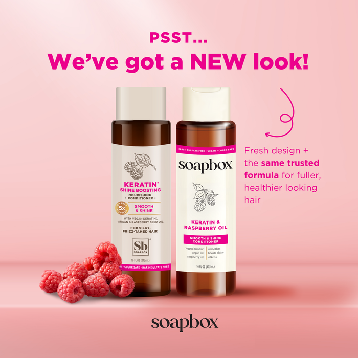Two bottles of Soapbox hair care with new packaging are displayed against a pink background. The left one features vegan keratin, the right boasts raspberry seed oil. Fresh raspberries are in front, with text highlighting a new look enriched with shine-boosting argan oil.