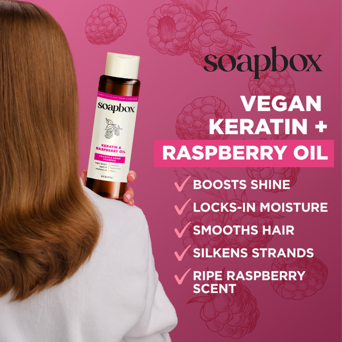 A long-haired person holds a bottle of Soapbox Vegan Keratin & Raspberry Seed Oil Smoothing Conditioner against a pink backdrop with raspberry illustrations. Benefits include vegan keratin for shine, moisture lock, smoothness, silkiness, and a ripe raspberry scent.