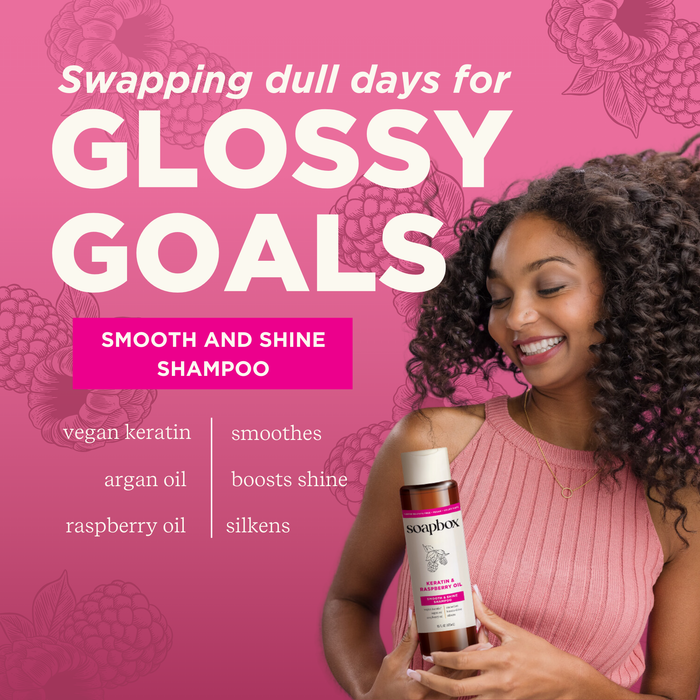 A smiling woman with curly hair holds a bottle of Soapbox Vegan Keratin & Raspberry Seed Oil Smoothing Shampoo. The pink backdrop with raspberries and floral designs highlights its benefits: vegan keratin, 5X vitamin complex, raspberry seed oil for smoothing shine and vibrant color-safe hair.