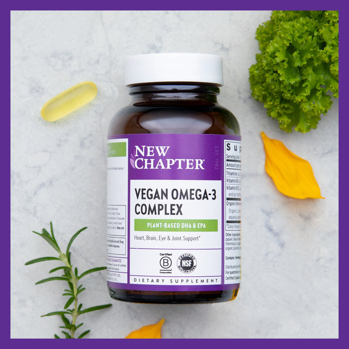 A bottle of New Chapter Vegan Omega-3 Complex, with pure algal oil, is surrounded by rosemary, a lettuce leaf, a yellow flower petal, and a capsule. The label showcases plant-based DHA & EPA for supporting heart, brain, eyes, and joints.