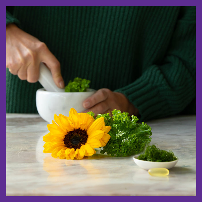 A person in a dark green sweater uses a mortar and pestle with parsley, embracing plant-based ingredients. Nearby are a sunflower, more parsley, and a dish with yellow liquid likely New Chapters Vegan Omega-3 Complex, all on marble. The image features a purple border.
