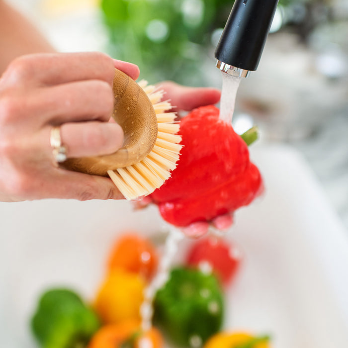 A person uses a Full Circle Veggie Brush to clean a red bell pepper under running water in the kitchen sink, while other colorful peppers sit in the background, ensuring freshness and completing the cycle of kitchen care.