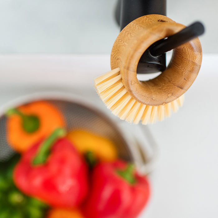 A Full Circle Veggie Brush with bristles hangs from a faucet above a stainless steel colander filled with red and yellow bell peppers, enhancing the clean, bright kitchen environment and serving as the perfect tool for produce cleaning among fresh ingredients.