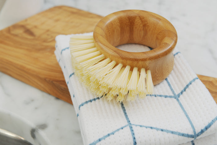 The Full Circle Veggie Brush, featuring recycled beige bristles and a bamboo handle, lies on a folded white dish towel with blue stripes on a light wooden cutting board atop a marble surface.