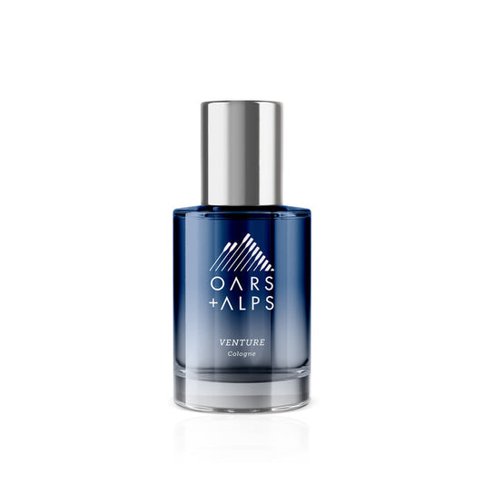 A blue glass bottle of Oars + Alps VENTURE Cologne with a silver cap and white logo is shown on a white background, reflecting the bergamot-inspired fresh marine and citrus fragrance of this signature scent.