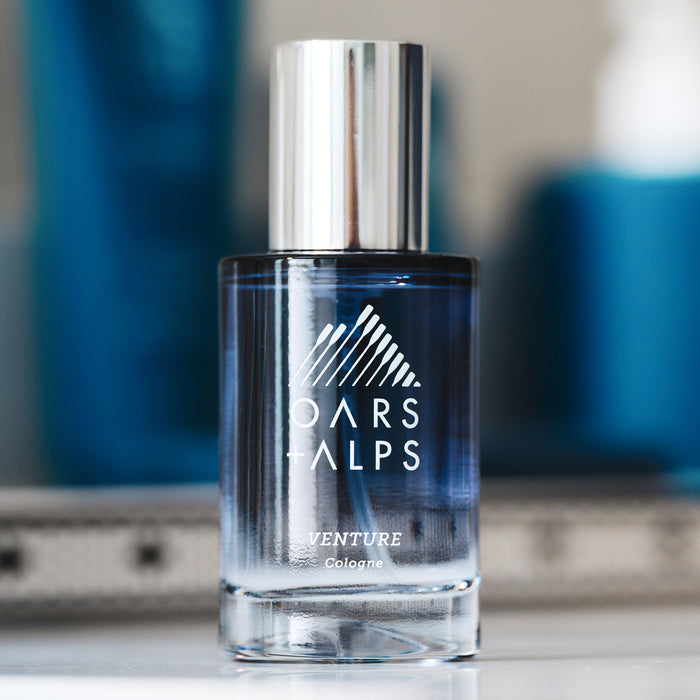 A clear glass bottle of Oars + Alps VENTURE Cologne with a silver cap and white branding sits on a reflective surface against a blurred blue background, evoking its fresh blend of marine and citrus notes.