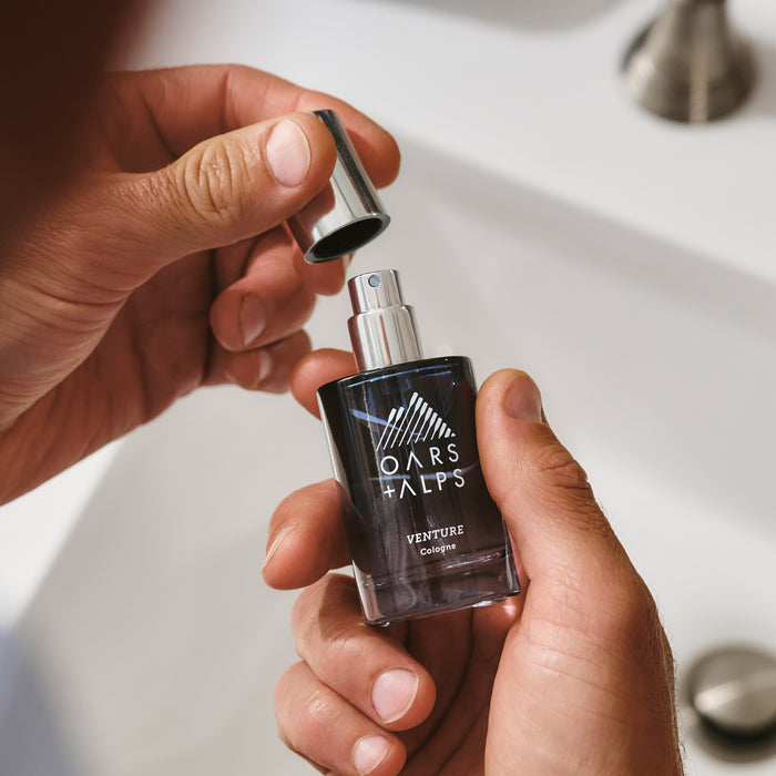 A person stands over a bathroom sink, gripping Oars + Alps VENTURE Cologne in one hand and removing the silver cap with the other, ready to experience its fresh marine and citrus scent.