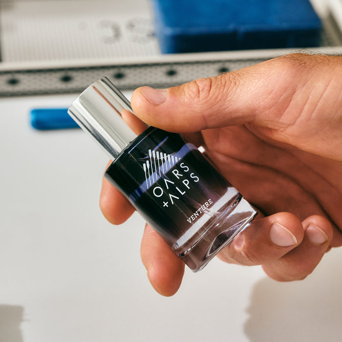 A hand holds a small bottle of Oars + Alps VENTURE Cologne with a silver cap on a white surface, highlighting its refreshing marine and citrus notes.