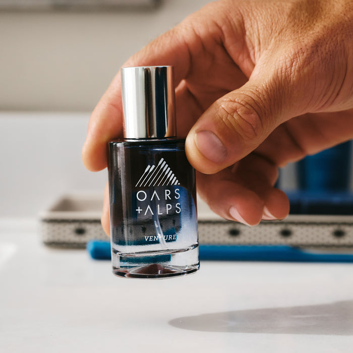 A hand holds a bottle of Oars + Alps VENTURE Cologne, featuring a silver cap and blue gradient glass, in front of a bathroom sink—showcasing its fresh marine and citrus scent.