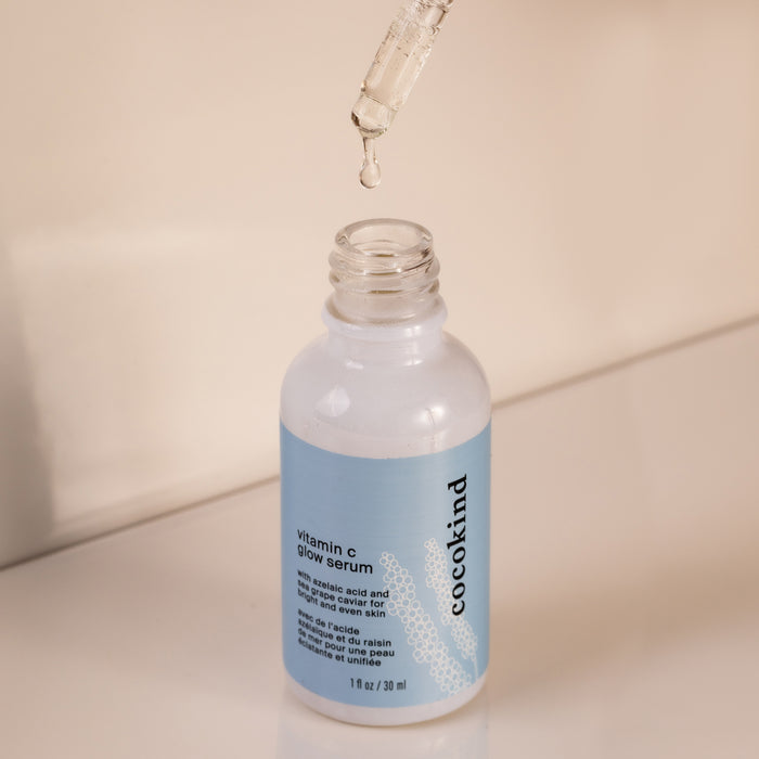 A dropper hovers over an open bottle of Cocokind Vitamin C Glow Serum with hyaluronic acid. The bottle, featuring a blue label and white text, rests gracefully on a smooth, reflective surface.