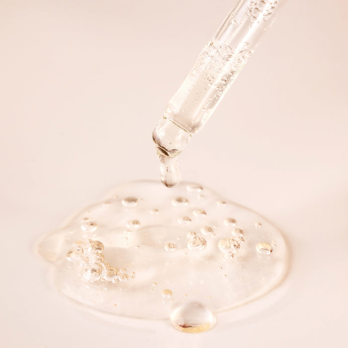A clear dropper releases a drop of Cocokinds Vitamin C Glow Serum onto a smooth surface. With small bubbles visible, some liquid pools below, highlighting the serums clarity and potency against a soft, neutral background.