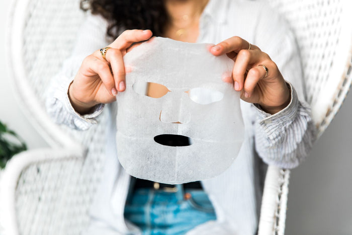 A person in a striped shirt sitting on a white wicker chair holds an ORGAID Vitamin C & Revitalizing Organic Sheet Mask made with Ecoderma fabric technology. Their face is partly hidden by the organic mask, and they have long, curly hair.