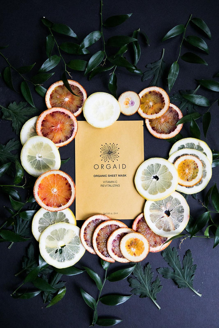 A packet of ORGAIDs Vitamin C & Revitalizing Organic Sheet Mask in vibrant yellow is encircled by lemon and blood orange slices, with green leaves enhancing the display against a dark background, showcasing the rejuvenating benefits of Ecoderma fabric technology.