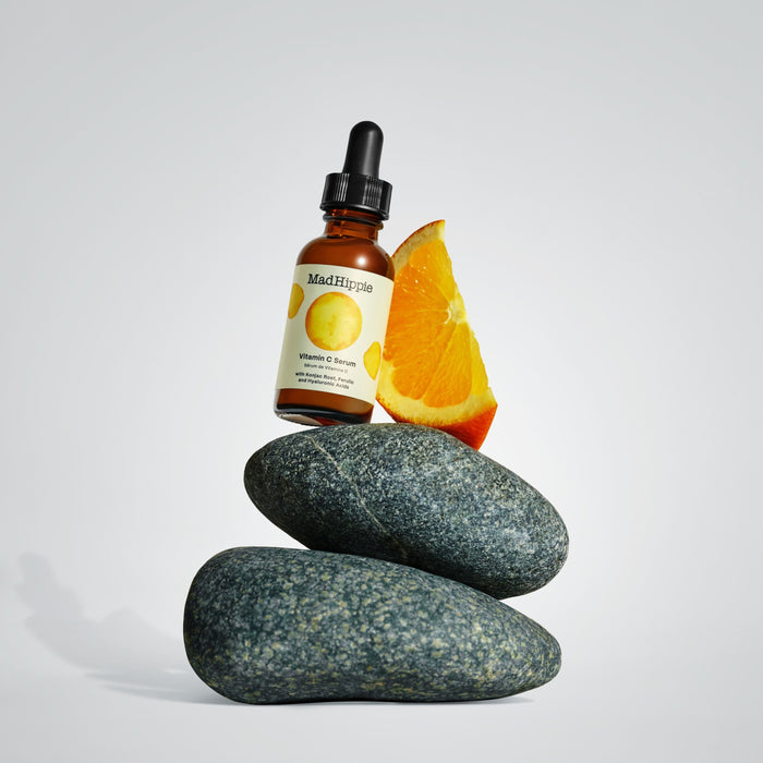 A dropper bottle of Mad Hippies Vitamin C Serum rests on three smooth gray stones, illustrating antioxidant power. A slice of orange emphasizes the citrus theme against a plain, light gray backdrop.