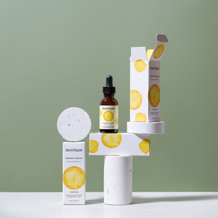 Mad Hippies Vitamin C Serum, rich in antioxidants, is displayed with two boxes on minimalist white cylinder stands against a light green background.