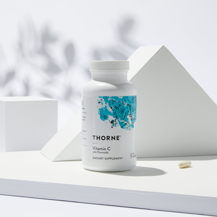 A Thorne Vitamin C With Flavonoids bottle, featuring a blue abstract design that highlights its antioxidant benefits, sits on a white geometric surface. A capsule lies nearby with soft shadows and a blurred leaf pattern in the background.