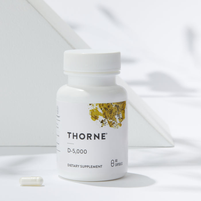 A white bottle labeled Thorne Vitamin D-5000 is showcased against a minimalist backdrop with geometric patterns. One Vitamin D3 capsule is in front, emphasizing its immune support benefits, with the scene brightly lit and soft shadows present.