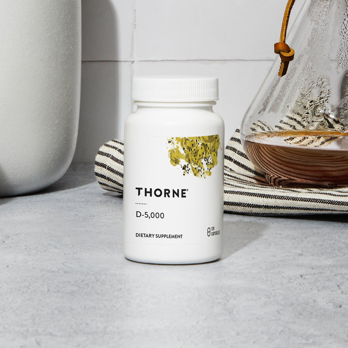A white bottle of Thorne Vitamin D-5000 sits on a gray countertop near a striped towel and a glass coffee pot with a leather tie, set against a white tiled wall.