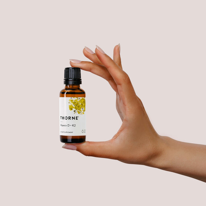 A hand gently cradles a small bottle labeled Thornes Vitamin D + K2, known for immune support. It has a black cap and a white label with floral design, set against a neutral beige backdrop.
