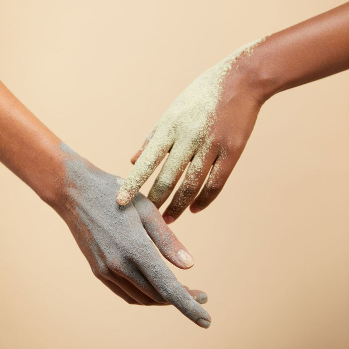 Two hands coated in vivid powders, light green and gray like volcanic ash, connect against a neutral backdrop. This mirrors the deep cleanse provided by Terra Beauty Bars Volcanic Ash Dry Facial Clay Mask made with vegan ingredients.