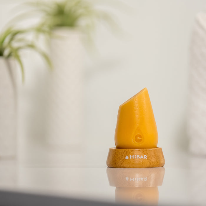 A solid orange HiBAR Volumize Conditioner Bar stands upright on a wooden base, ideal for adding volume to fine hair. The reflective surface enhances its sheen while blurred potted plants add greenery in the background. The base showcases the HiBAR logo.