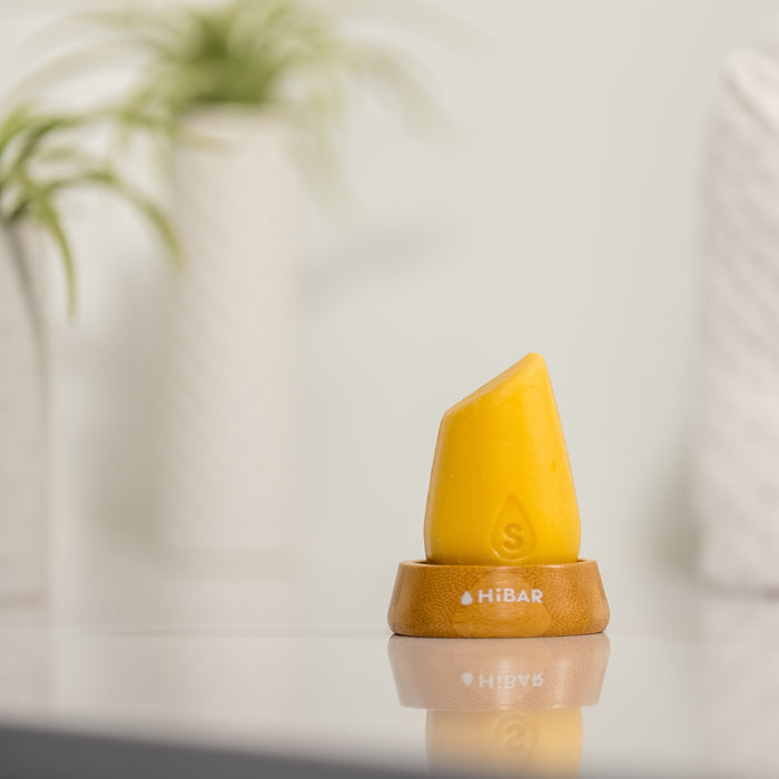 A yellow Volumize Shampoo by HiBAR stands upright on a wooden base, ideal for thin hair. The unique-shaped bar is set against a backdrop of blurred green plants in white pots, enhancing a clean and minimalistic atmosphere.