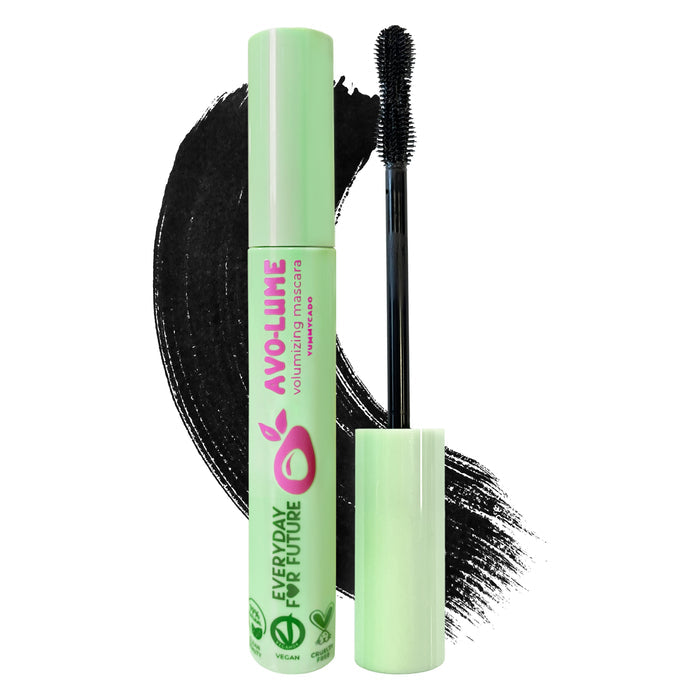 A tube of Everyday For Futures Volumizing Avocado Mascara, with sustainable packaging and a green cap, sits beside a halved avocado. The brush extends outside, and black mascara streaks are smeared in the background.