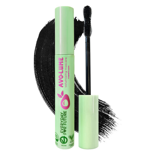 A tube of Everyday For Futures Volumizing Avocado Mascara, with sustainable packaging and a green cap, sits beside a halved avocado. The brush extends outside, and black mascara streaks are smeared in the background.