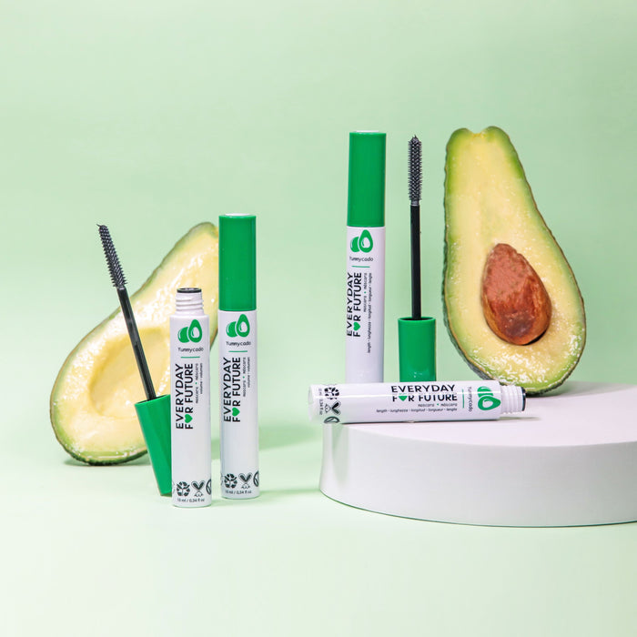 Several tubes of Everyday For Futures Volumizing Avocado Mascara with Castor Oil, featuring green caps, are displayed next to a halved avocado on a white round platform against a light green background. Some tubes are open with brushes shown, and the eco-friendly packaging promotes vegan and cruelty-free lash growth.