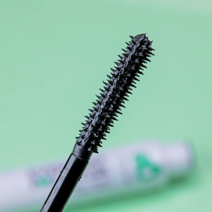 A close-up of the Volumizing Avocado Mascara wand from Everyday For Future features short, spiky bristles coated in black mascara. The blurred green background suggests its vegan, cruelty-free packaging, emphasizing its commitment to lash growth with nourishing castor oil.