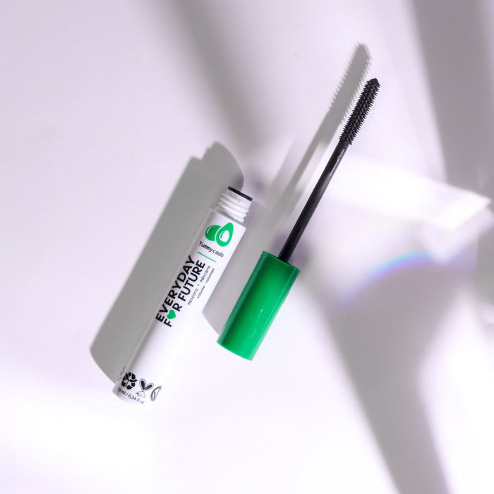 A white Volumizing Avocado Mascara with Castor Oil tube from Everyday For Future, displaying black text, a green cap, and brush wand sits on a white surface. Sunlight casts a rainbow and soft shadows, emphasizing its vegan and cruelty-free promise for lash growth.