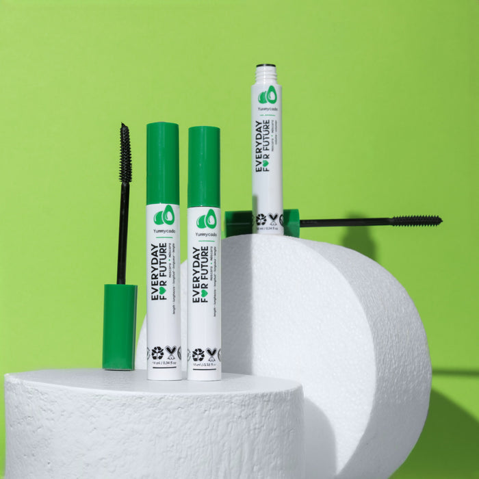 Two tubes of Everyday For Futures Volumizing Avocado Mascara with Castor Oil and green caps are elegantly placed on white cylindrical platforms against a vibrant green background. One tube is open, showcasing the brush wand. The vegan and cruelty-free symbols emphasize its ethical beauty commitment.
