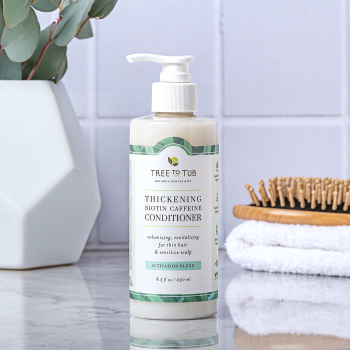 A bottle of Tree To Tub Volumizing Caffeine & Biotin Conditioner for Fuller, Thicker Hair with a pump sits on a bathroom counter. Behind it are a hairbrush, folded white towels, and a vase with eucalyptus leaves against pristine white tiles.