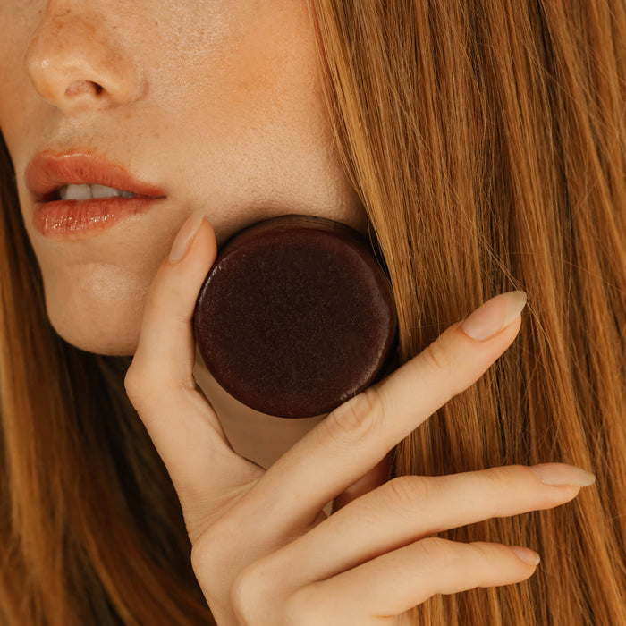 A woman with long red hair holds the Good Juju Body & Home Volumizing Conditioner Bar for Oily/Fine Hair near her cheek, showcasing her natural makeup and nude nails. Her lips are slightly parted and only part of her face is visible.