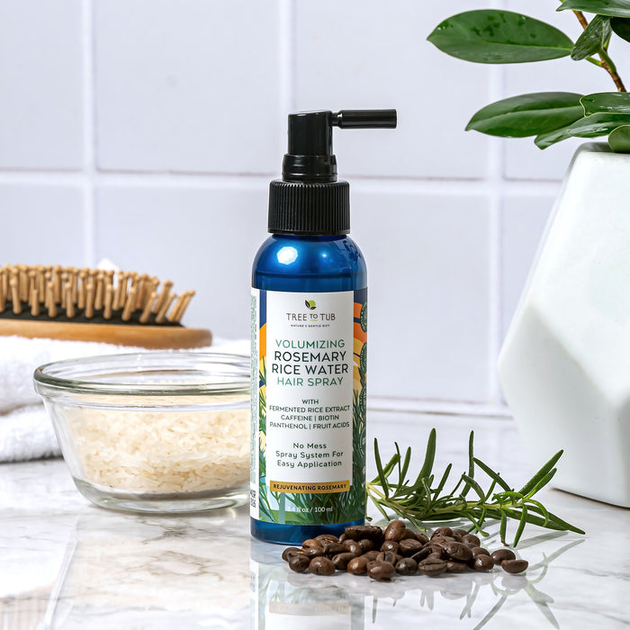 A bottle of Tree To Tubs Volumizing Rosemary & Rice Water Hair Spray for Fuller, Thicker Hair rests on a white counter. Nearby are a bowl of rice, coffee beans, a hairbrush, and plant leaves against white tiled walls, crafting an elegant hair care setting.