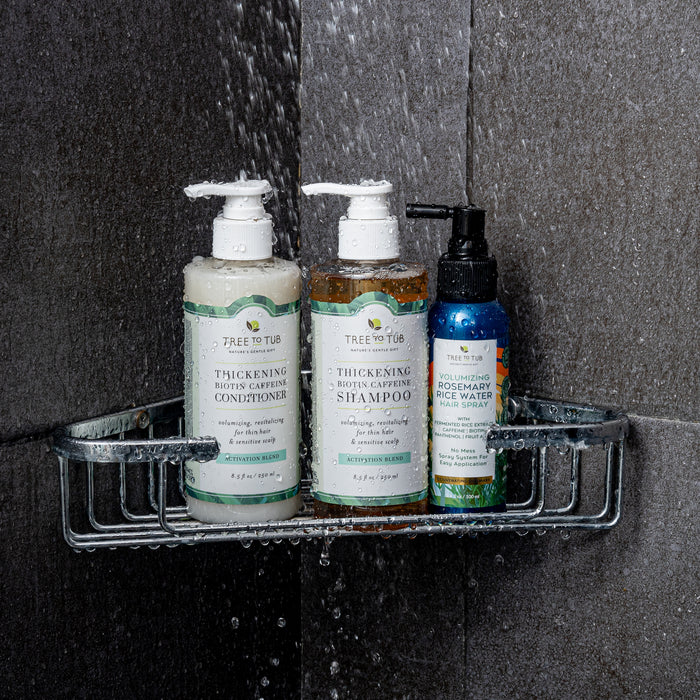 A Tree To Tub shower caddy showcases your hair care trio: a conditioner, shampoo, and Volumizing Rosemary & Rice Water Hair Spray for fuller hair. The bottles glisten with droplets against dark gray tiles.
