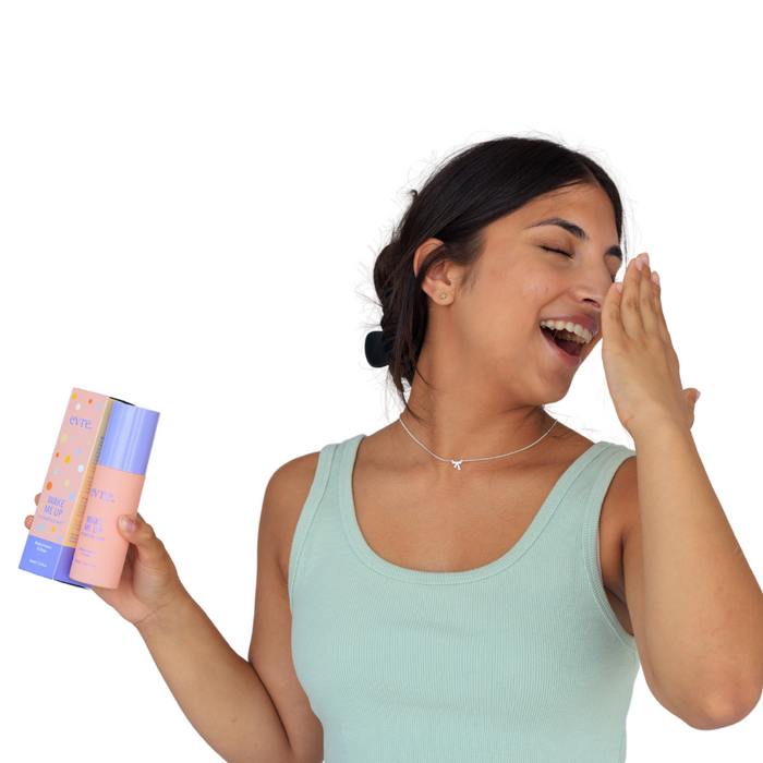 A woman with dark hair tied back, wearing a necklace and green tank top, smiles with closed eyes. Her left hand near her face holds an evre. Wake Me Up Watermelon & Rose Face Mist and its box against a white background.