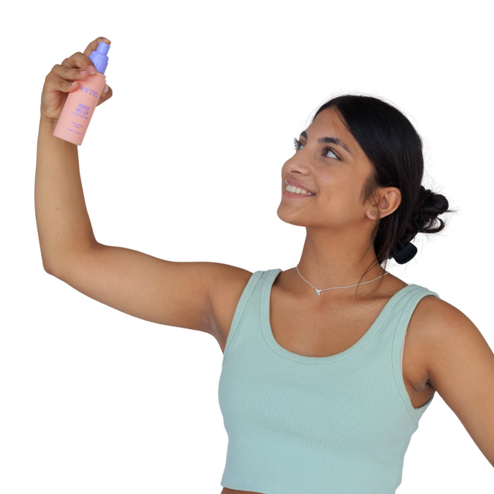 Wearing a light green tank top, someone holds up a bottle of evre.s Wake Me Up Watermelon & Rose Face Mist, smiling and looking upwards. The peach bottle with a purple cap stands out against the plain white background.