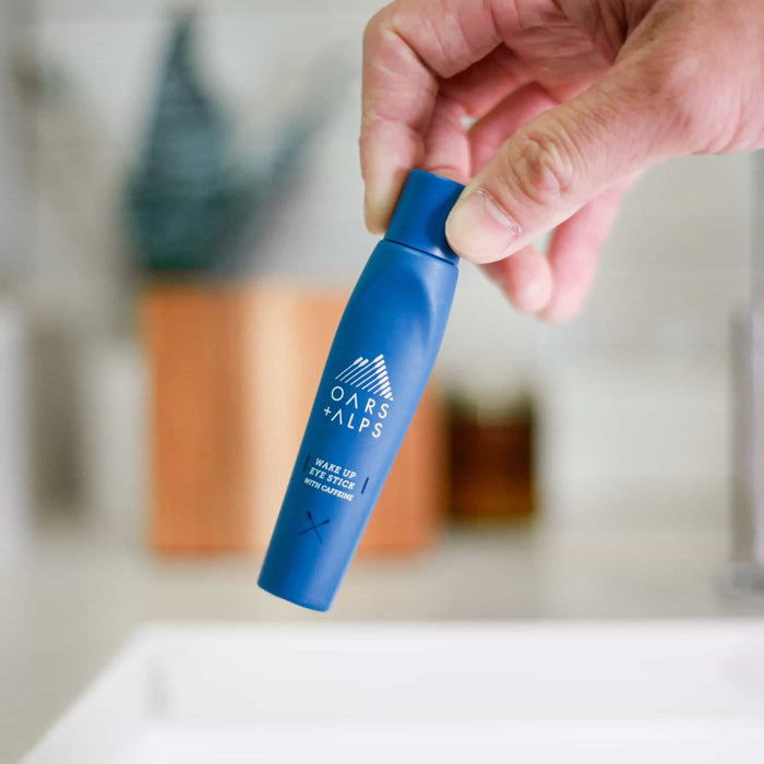 A hand holds an Oars + Alps Wake Up Eye Stick—an eye serum with caffeine to refresh puffy under-eyes—above a bathroom sink with a softly blurred background.