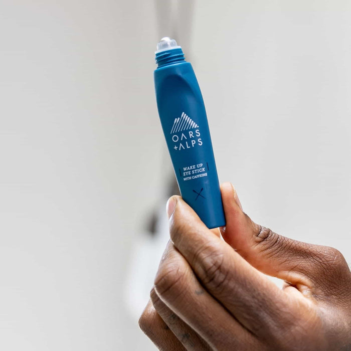 A hand holds an open Oars + Alps Wake Up Eye Stick, revealing its rollerball applicator—formulated with Caffeine and Aloe Vera—against a softly blurred background.