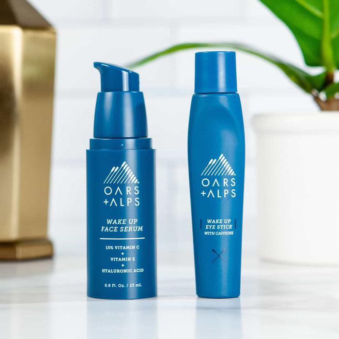 Oars + Alps Wake Up Eye Stick stands on a white surface, with a gold lamp and green plant blurred in the background.