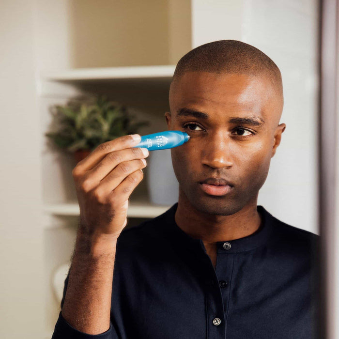 A man in a dark shirt stands before a mirror applying Oars + Alps Wake Up Eye Stick with caffeine under his eye using a blue applicator. A plant on the shelf in the background highlights his focus on reducing puffiness.